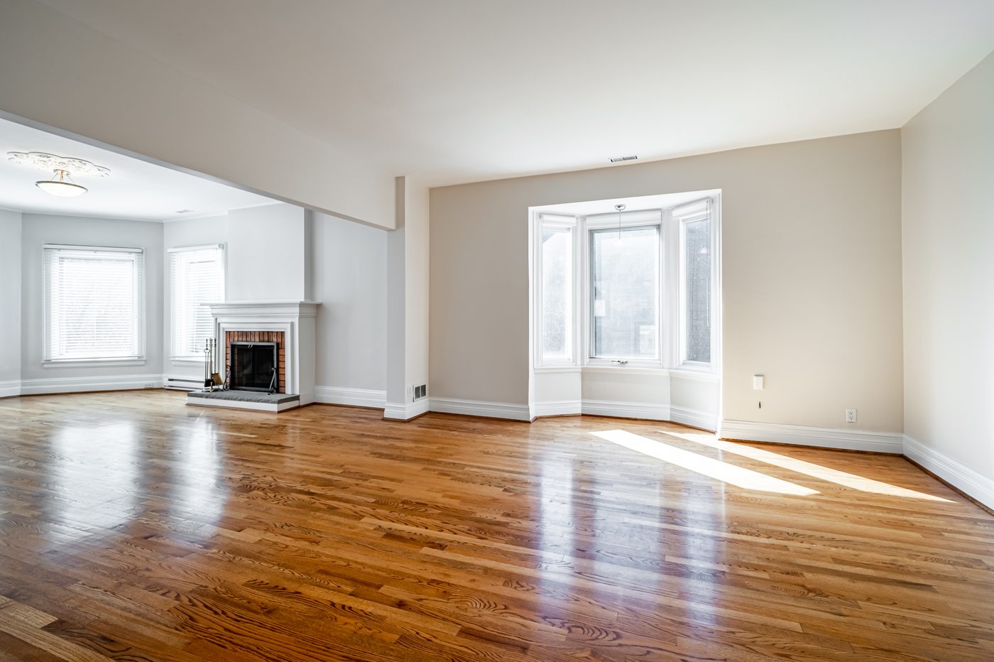 Original photograph of a vacant living and dining room with hardwood floors, a brick fireplace, and a bay window — unstaged and untouched